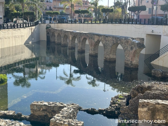 Tramo del acueducto romano de La Carrera parcialmente sumergido en agua estancada, con reflejos de palmeras y edificios urbanos al fondo en Almuñécar.