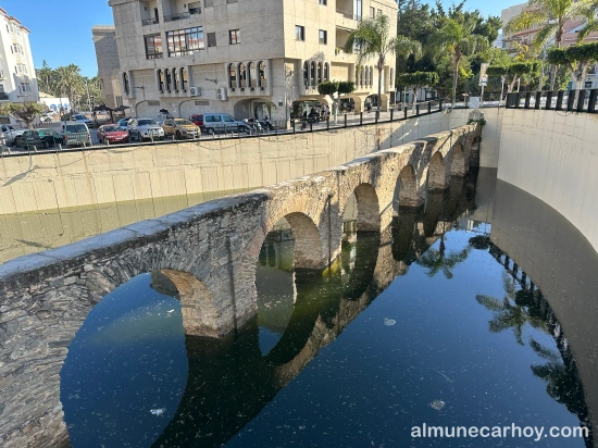 Tramo del acueducto romano de La Carrera parcialmente sumergido en agua estancada, con los arcos reflejados en la lámina de agua y edificios y palmeras al fondo en Almuñécar.