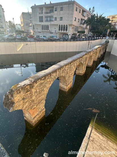 Tramo del acueducto romano de La Carrera parcialmente sumergido en agua estancada, con edificios residenciales y barandilla perimetral al fondo en Almuñécar.