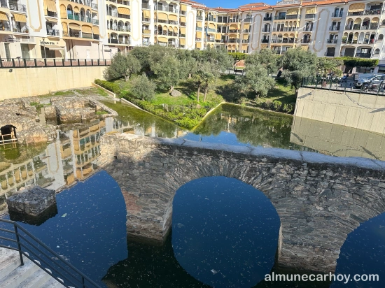 Tramo del acueducto romano de La Carrera rodeado por agua estancada, con zona ajardinada con olivos y edificios residenciales al fondo en Almuñécar.