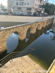 Tramo del acueducto romano de La Carrera parcialmente inundado, con agua estancada junto a las escaleras de acceso y edificios al fondo en Almuñécar.