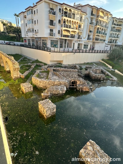 Restos de las termas romanas de La Carrera parcialmente cubiertos por agua estancada, con bloques de piedra emergiendo y edificios residenciales al fondo en Almuñécar.