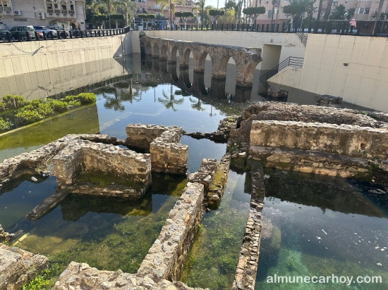 Restos de las termas romanas de La Carrera parcialmente cubiertos por agua estancada, con el tramo del acueducto romano al fondo y edificios urbanos alrededor en Almuñécar.