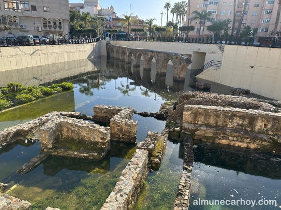 Restos de las termas romanas de La Carrera parcialmente cubiertos por agua estancada, con el tramo del acueducto romano al fondo y edificios urbanos alrededor en Almuñécar.